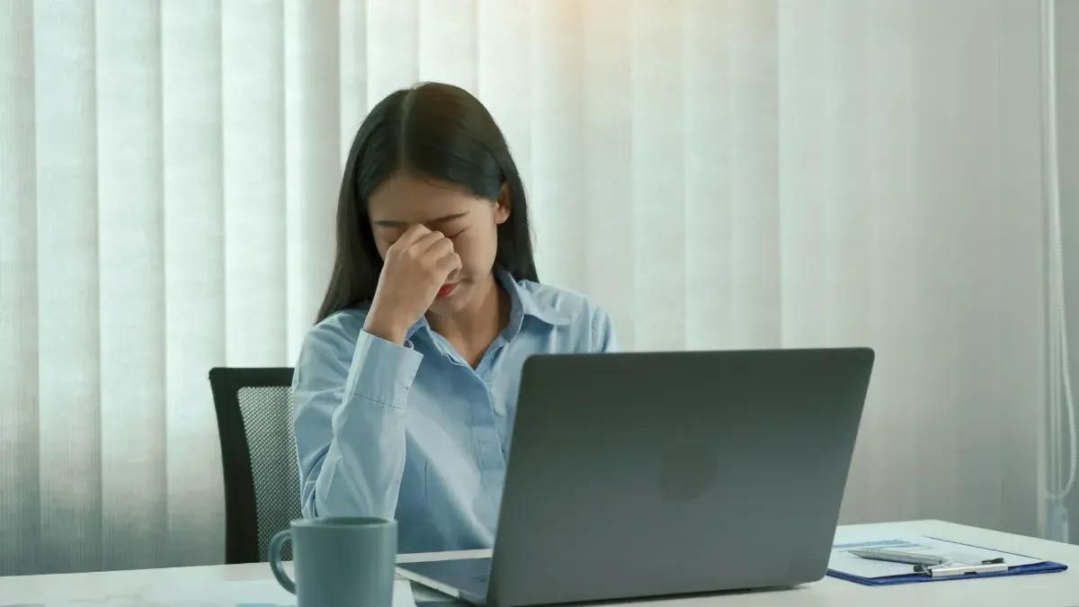 淺藍襯衫女性坐在筆電前，低頭捏著眉心。