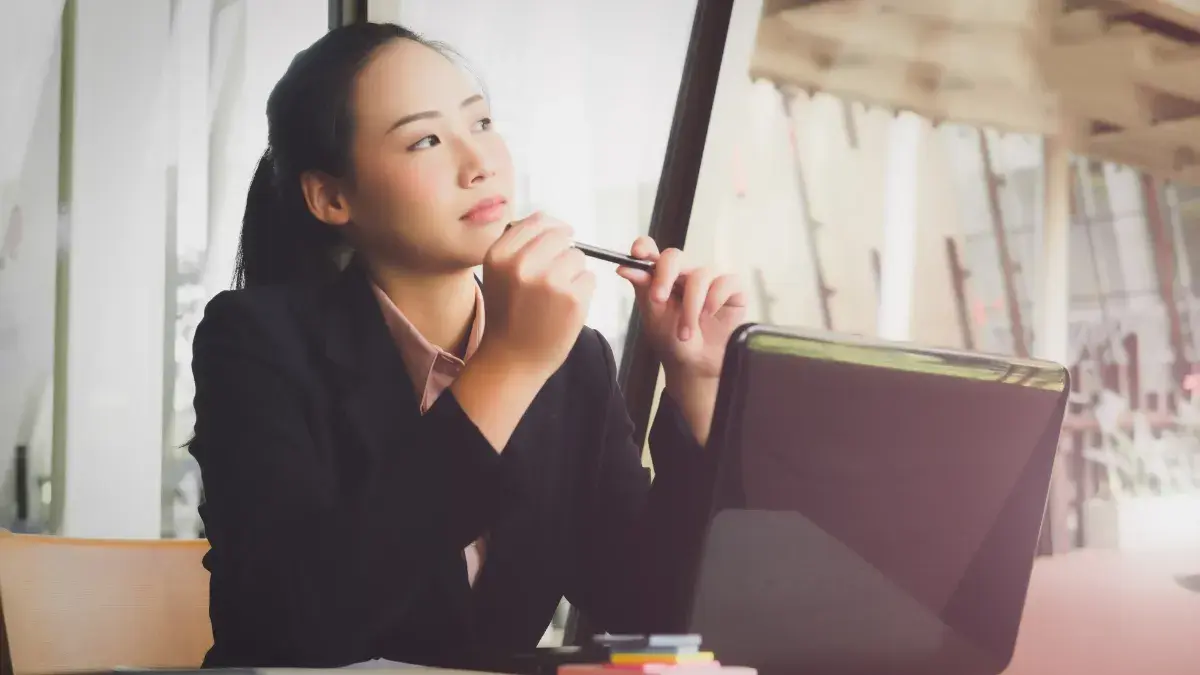 深色上衣女性坐在筆電旁,拿著筆思考。